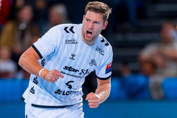 Ein männlicher Handballspieler in einem weißen Trikot ballt während eines Spiels die Faust und schreit in Jubel aus, während im Hintergrund unscharfe Zuschauer zu sehen sind.