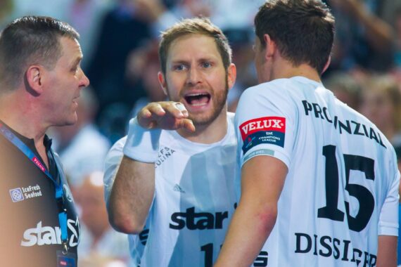 Drei männliche Handballspieler in weißen Trikots, einer gestikuliert und spricht intensiv, möglicherweise in einer Diskussion oder einem Streit, während ein anderer Spieler und ein Trainer oder Schiedsrichter zuhören. Die Szene scheint während eines Spiels zu spielen.