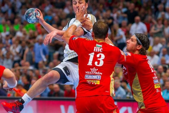 Ein Handballspieler in einem weißen Trikot springt zum Wurf, während zwei Verteidiger in roten Trikots ihn abwehren. Das Spiel findet in einer überfüllten Halle mit einer unscharfen Menschenmenge im Hintergrund statt.
