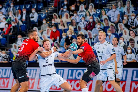 Zwei Handballspieler in roten Trikots blockieren einen Spieler in einem weißen Trikot, der den Ball während eines Spiels hält; maskierte Zuschauer und Pappausschnitte füllen die Zuschauerplätze im Hintergrund.