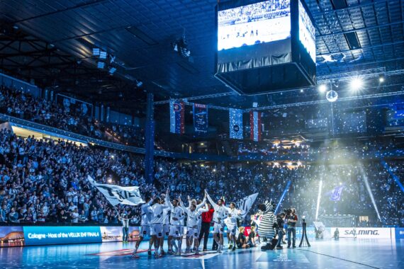 Eine Handballmannschaft feiert auf einem hell erleuchteten Hallenplatz, winkt mit Fahnen und lässt Konfetti fallen. Eine große Menschenmenge füllt die Plätze in der Halle, und über dem Spielfeld hängt eine große Leinwand, auf der die Feierlichkeiten zu sehen sind.