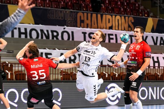Ein Handballspieler in einem weißen Trikot springt in die Luft, um den Ball zu werfen, während zwei gegnerische Spieler in roten Trikots versuchen, ihn während eines Hallenspiels zu blockieren. Im Hintergrund sind Zuschauersitze und Banner zu sehen.