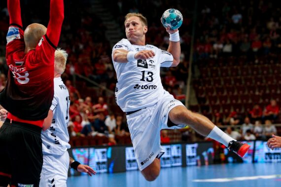 Ein männlicher Handballspieler in einem weißen Trikot springt in die Luft, um einen Schuss abzugeben, wobei er den Ball in der rechten Hand hält, während ein Verteidiger in Rot versucht, ihn abzuwehren. Die Zuschauer schauen von der Tribüne im Hintergrund zu.