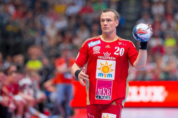 Ein männlicher Handballspieler in einem roten Trikot mit der Nummer 20 hält einen Ball und bereitet sich auf den Wurf während eines Spiels vor. Der Hintergrund zeigt eine Reithalle mit einer unscharfen Menschenmenge.