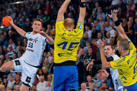 Ein Handballspieler in einem weißen Trikot springt, um den Ball zu schießen, während drei Verteidiger in gelben Trikots die Arme heben, um ihn abzublocken, während im Hintergrund eine große Menschenmenge zusieht.