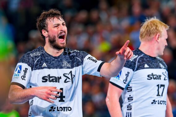 Ein männlicher Handballspieler in einem weiß-schwarzen Trikot schreit während eines Spiels leidenschaftlich und gestikuliert mit der Hand, während ein anderer Spieler neben ihm auf einem hell erleuchteten Hallenplatz steht.