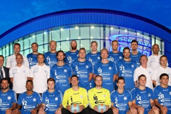 Eine Herrenhandballmannschaft in blauen Trikots und Trainer in weißen Hemden posieren für ein Gruppenfoto vor einem modernen Gebäude mit Glasscheiben und einem Schild mit der Aufschrift SCHWALBE.