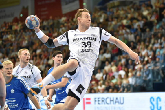 Ein Handballspieler in einem weißen Trikot springt in der Luft und bereitet sich darauf vor, den Ball während eines Spiels in Richtung Tor zu werfen, während Spieler in blauen Trikots und die Zuschauer im Hintergrund aufmerksam zusehen.