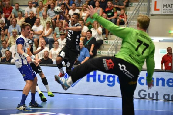 Ein Handballspieler in einem schwarzen Trikot springt, um den Ball zu schießen, während ein Torwart in Grün und zwei Verteidiger in Weiß sich darauf vorbereiten, den Ball zu blocken. Das Spiel findet in einer überfüllten Halle mit Zuschauern statt.