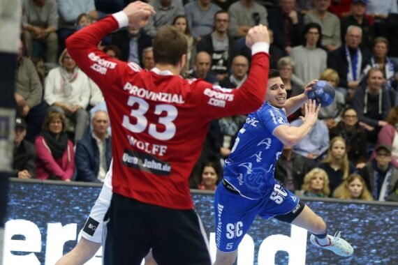 Ein Handballspieler in einem blauen Trikot springt auf, um den Ball zu schießen, während ein Torwart in einem roten Trikot mit der Nummer 33 die Arme zum Abwehren hochreißt.
