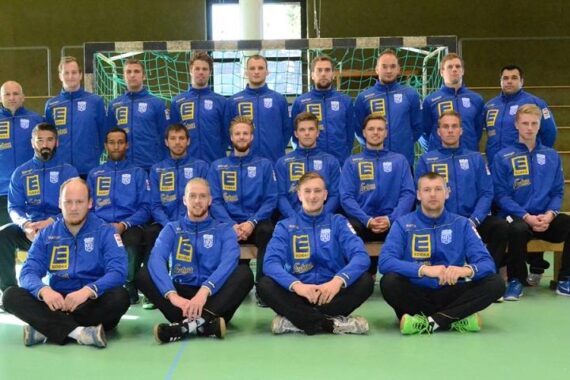 Ein Herrensportteam in blauen Jacken posiert für ein Gruppenfoto in einer Sporthalle, sitzend und stehend in drei Reihen vor einem Tornetz.