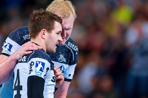 Zwei männliche Handballspieler in schwarz-weißen Trikots stehen dicht beieinander, der eine hat den Arm um die Schulter des anderen gelegt, und es scheint, als würden sie sich während eines Spiels unterhalten oder sich gegenseitig unterstützen. Der Hintergrund ist mit Zuschauern unscharf.