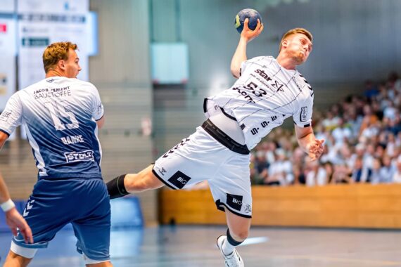 Ein Handballspieler in einem weißen Trikot springt durch die Luft, um ein Tor zu erzielen, während zwei gegnerische Spieler in Blau versuchen, ihn zu blockieren. Eine Menschenmenge verfolgt das intensive Spiel von der Tribüne aus in einer hell erleuchteten Halle.
