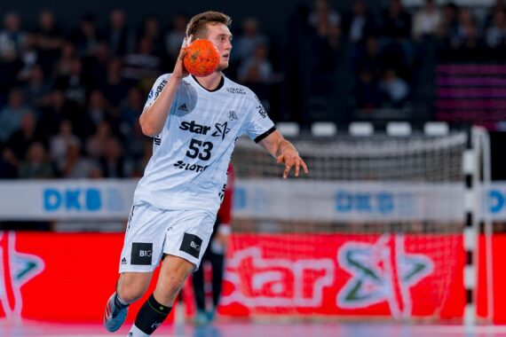 Ein männlicher Handballspieler in einem weißen Trikot mit der Nummer 53 bereitet sich darauf vor, während eines Spiels einen orangefarbenen Ball zu werfen, während im Hintergrund ein Tor und Zuschauer zu sehen sind.