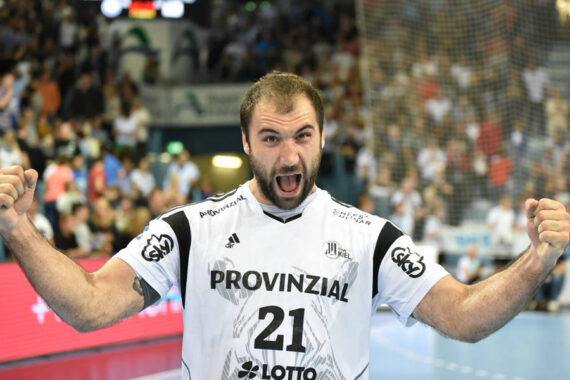 Ein männlicher Handballspieler in einem weißen Trikot mit der Nummer 21 jubelt mit erhobenen Fäusten und offenem Mund auf einem Spielfeld, mit einer unscharfen Menschenmenge im Hintergrund.