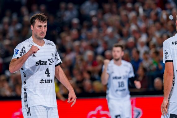 Ein männlicher Handballspieler in einem weißen Trikot mit der Nummer 4 steht auf dem Spielfeld, hinter ihm zwei Mannschaftskameraden in ähnlichen Trikots und eine verschwommene Menschenmenge im Hintergrund.