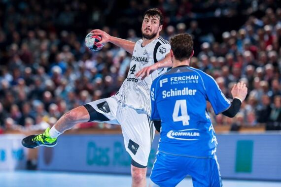Ein Handballspieler in einem weißen Trikot springt mit einem Ball und versucht zu schießen, während ein Verteidiger in einem blauen Trikot mit der Schindler 4 auf dem Rücken ihn blockiert. Die Aktion findet in einer überfüllten Halle statt.