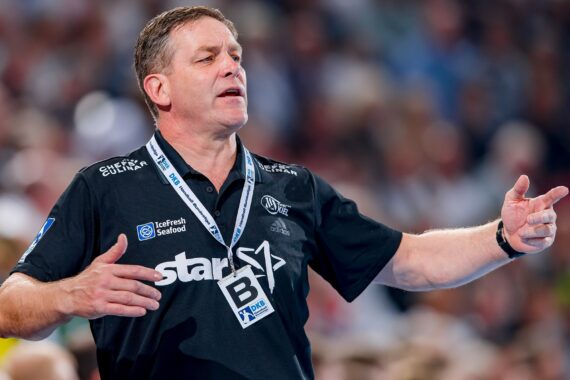 Ein Sporttrainer in einem schwarzen Polohemd gestikuliert mit beiden Händen, als würde er während eines Spiels Anweisungen geben. Er trägt ein Schlüsselband und steht vor einer verschwommenen Menschenmenge.