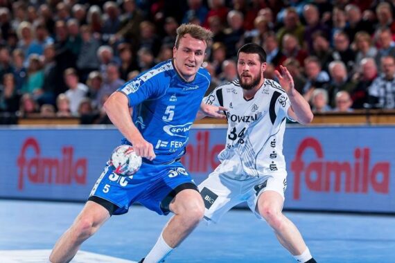 Zwei männliche Handballspieler in blau-weißen Trikots kämpfen auf einem Hallenplatz um den Ball, während im Hintergrund eine Zuschauermenge von der Tribüne aus zusieht.