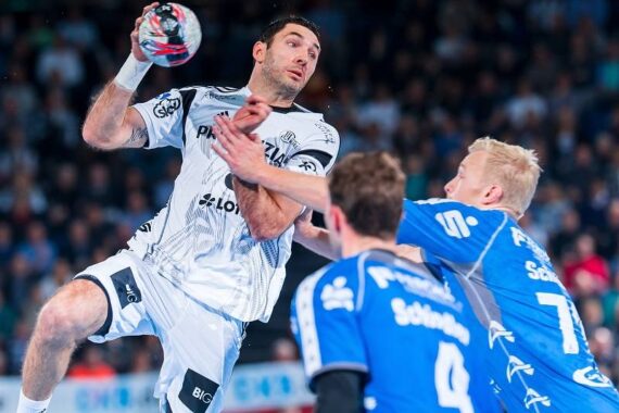 Ein Handballspieler in einem weißen Trikot springt, um den Ball zu werfen, während zwei gegnerische Spieler in Blau versuchen, ihn während eines Spiels zu verteidigen. Das Spiel findet auf einem Hallenplatz statt, mit einer unscharfen Zuschauermenge im Hintergrund.