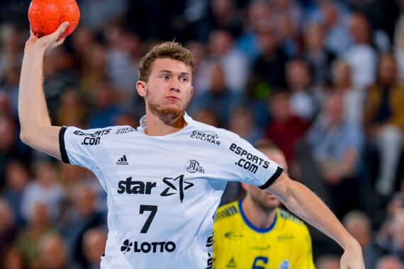 Ein männlicher Handballspieler in einem weißen Trikot, der einen orangefarbenen Ball in der rechten Hand hält, bereitet sich auf den Wurf vor. Er befindet sich mitten in der Aktion auf einem Hallenplatz, mit einem anderen Spieler und einer unscharfen Menge im Hintergrund.