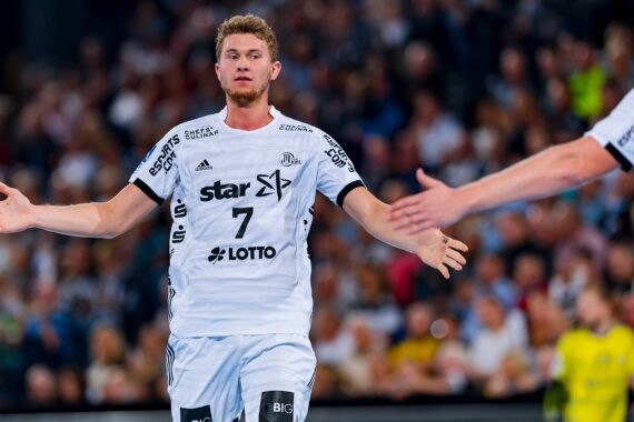 Ein männlicher Handballspieler in einem weißen Trikot mit der Nummer 7 steht mit ausgestreckten Armen da und scheint zum Abklatschen bereit zu sein, während ein anderer Spieler nach ihm greift. Eine verschwommene Menschenmenge schaut im Hintergrund zu.