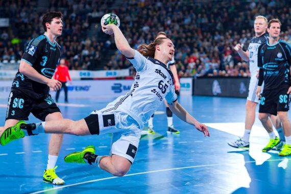 Ein Handballspieler in einem weißen Trikot springt in der Luft, um den Ball in Richtung Tor zu werfen, während gegnerische Spieler in schwarzen Trikots den Ball verteidigen, auf einem Hallenplatz mit einer Menschenmenge im Hintergrund.