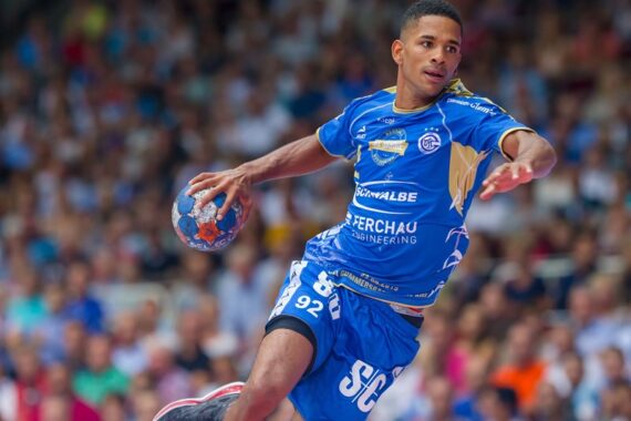 Ein männlicher Handballspieler in einem blauen Trikot springt mit einem Ball in der Hand in die Luft und bereitet sich darauf vor, ihn zu werfen. Im Hintergrund der Halle ist eine verschwommene Menschenmenge zu sehen.