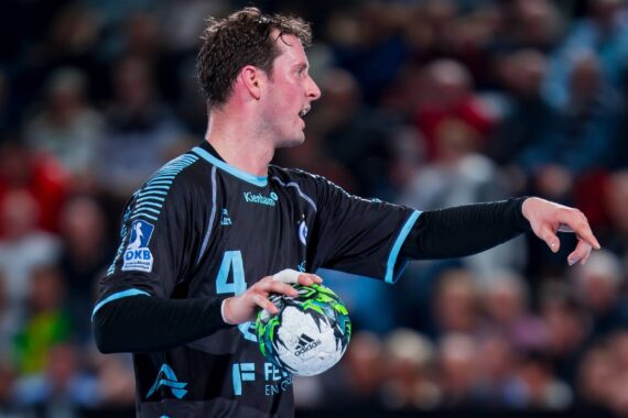 Ein männlicher Handballspieler in einem schwarzen Trikot hält einen Handball in der linken Hand und gestikuliert mit der rechten Hand, konzentriert während eines Spiels mit einer unscharfen Menschenmenge im Hintergrund.