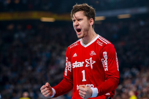 Ein männlicher Handballspieler in einem roten Trikot mit der Nummer 1 ballt während eines Spiels die Fäuste und jubelt, während im Hintergrund eine verschwommene Menschenmenge zu sehen ist.