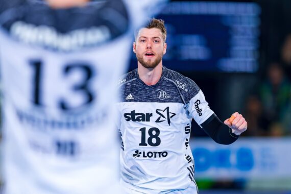 Ein männlicher Handballspieler in einem weiß-blauen Trikot mit der Nummer 18 ist auf dem Spielfeld in Aktion, während ein anderer Spieler in einem ähnlichen Trikot mit der Nummer 13 teilweise im Vordergrund zu sehen ist.