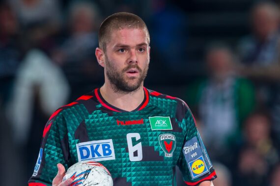 Ein männlicher Handballspieler in einem grün-schwarzen Trikot mit der Nummer 6 hält einen Ball und schaut konzentriert. Der Hintergrund ist unscharf und zeigt eine Sporthalle mit Zuschauern.