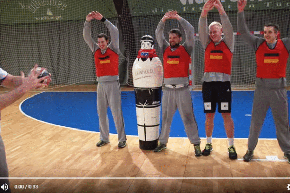 Vier Männer in roten Trainingswesten stehen mit erhobenen Armen neben einer gepolsterten Sportpuppe auf einem Hallenplatz, während ein anderer Mann einen Ball in der Hand hält und sich zum Wurf vorbereitet.
