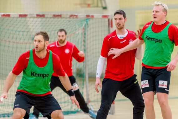 Vier männliche Sportler in roten Hemden und grünen Lätzchen spielen Hallenhandball; einer steht als Torwart vor dem Netz, während drei andere im Vordergrund aktiv spielen.