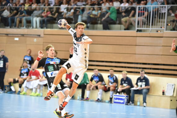 Ein Handballspieler in einem weißen Trikot springt mit dem Ball, um ein Tor zu erzielen, während ein Verteidiger in einem schwarz-grünen Trikot versucht, das Tor zu verhindern. Mitspieler, Gegner und Zuschauer schauen aus dem Hintergrund zu.