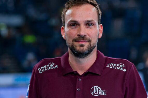 Ein Mann in einem kastanienbraunen Poloshirt mit den Logos "THW KIEL" und "CHEFS CULINAR" steht vor einem unscharfen Innenhintergrund vor der Kamera. Er hat kurze Haare und einen gestutzten Bart.