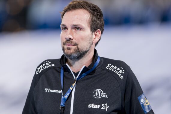 Ein Mann, der eine schwarze Sportjacke mit Logos trägt, steht in einem Haus und schaut nachdenklich. Er hat kurzes braunes Haar, einen gestutzten Bart und trägt ein Schlüsselband um den Hals. Der Hintergrund ist unscharf.