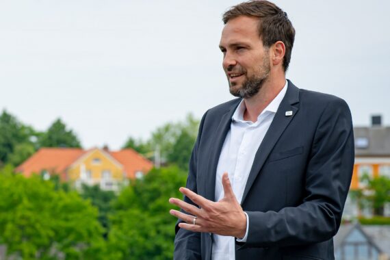 Ein Mann im Anzug spricht im Freien und gestikuliert mit einer Hand. Bunte Häuser und grüne Bäume sind im Hintergrund unter einem bewölkten Himmel verschwommen.