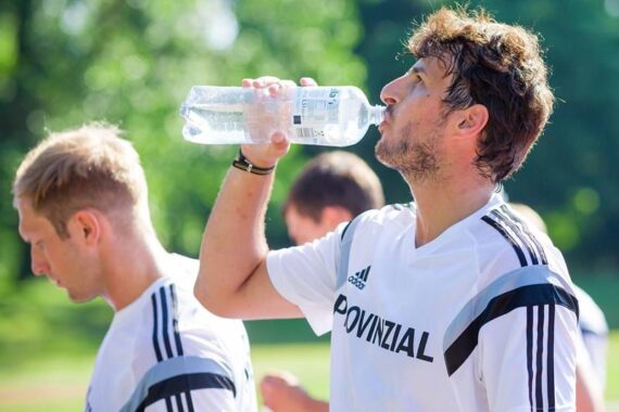 Zwei Männer in weißen Sporthemden mit der Aufschrift PROVINZIAL stehen draußen. Einer trinkt aus einer Wasserflasche, während der andere nach unten schaut. Der Hintergrund ist grün und sonnig, was auf ein Sporttraining hindeutet.
