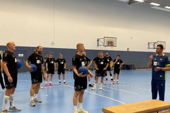 Eine Gruppe von Männern in Sportkleidung steht in einer Turnhalle, einige halten blaue Medizinbälle, während ein Trainer in Blau zu ihnen spricht. Im Hintergrund sind Basketballkörbe und Bänke zu sehen.