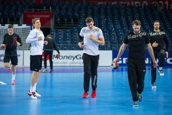 Fünf männliche Handballspieler wärmen sich in Sportkleidung auf einem Hallenplatz auf. Zwei von ihnen halten Handbälle in der Hand. Die Plätze in der Halle im Hintergrund sind größtenteils leer.