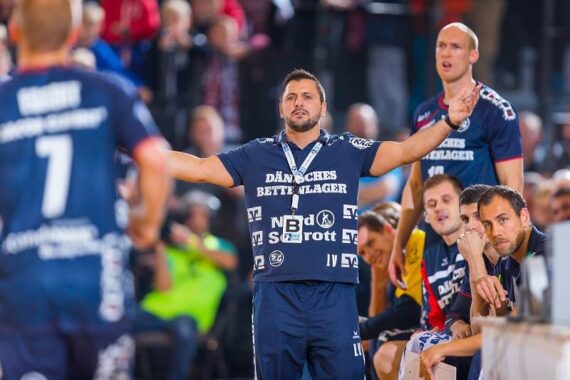Ein Handballtrainer in einem marineblauen Hemd gestikuliert während eines Spiels leidenschaftlich mit ausgestreckten Armen, während Spieler und Betreuer auf der Bank sitzen und aufmerksam zusehen. Im Hintergrund ist eine unscharfe Menschenmenge zu sehen.