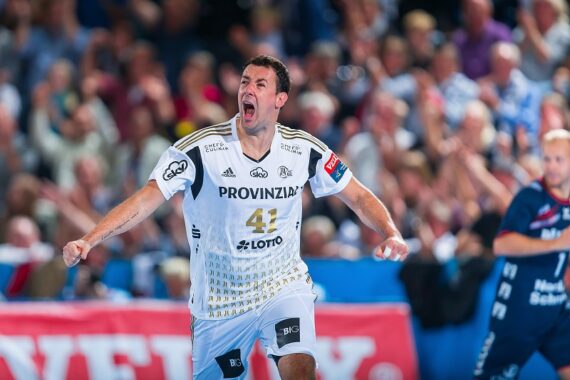 Ein männlicher Handballspieler in einem weißen Trikot jubelt mit ausgestreckten Armen auf einem Hallenplatz, umgeben von einer jubelnden Menge und anderen Spielern im Hintergrund.