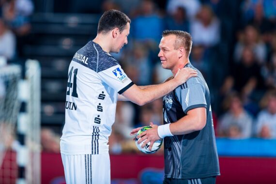 Zwei männliche Handballspieler stehen sich auf dem Spielfeld gegenüber. Ein Spieler in Weiß hat seinen Arm auf die Schulter des anderen gelegt, während der Spieler in Grau den Ball hält und lächelt. Im Hintergrund ist ein unscharfes Publikum zu sehen.