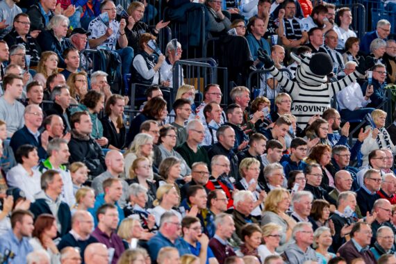 Eine große Menge von Zuschauern sitzt dicht gedrängt auf den Sitzen im Stadion und verfolgt eine Veranstaltung. Unter ihnen sticht eine Person in einem schwarz-weiß gestreiften Kostüm hervor und hebt die Arme.