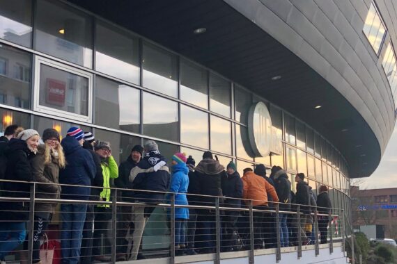 Eine lange Schlange von Menschen in Winterkleidung wartet an einem kalten Tag vor einem modernen Gebäude mit großen Fenstern. Einige Menschen stehen lächelnd und plaudernd an einem Metallgeländer.