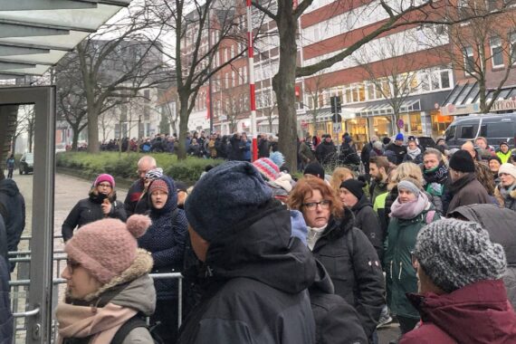 Eine große Menschenmenge in Winterkleidung wartet in einer langen Schlange draußen auf einer Stadtstraße. Einige Menschen tragen Mützen und Schals, und die Atmosphäre scheint kalt und bedeckt zu sein. Im Hintergrund sind Gebäude und kahle Bäume zu sehen.