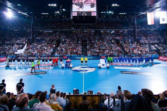 Eine große, mit Zuschauern gefüllte Halle zeigt zwei Handballmannschaften, die sich vor einem Spiel auf beiden Seiten des Spielfelds aufstellen, mit Offiziellen und Bannern in der Mitte.