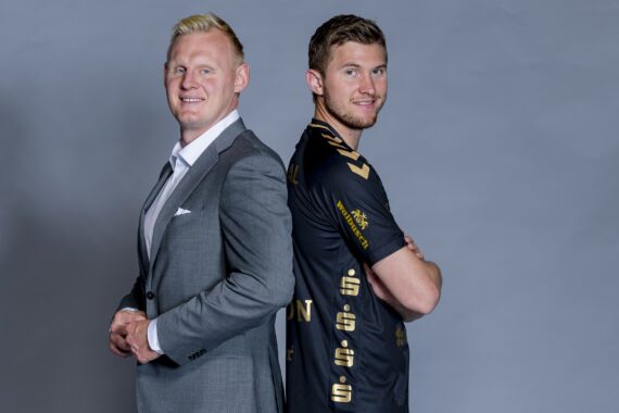 Zwei Männer stehen Rücken an Rücken. Der eine trägt einen grauen Anzug und lächelt, der andere trägt ein schwarz-goldenes Sporttrikot und hat die Arme verschränkt, beide stehen seitlich vor einem schlichten grauen Hintergrund.