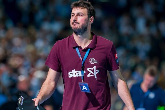 Ein Mann in einem kastanienbraunen Polohemd mit Stern und anderen Logos geht in einer Sportarena und hält ein Klemmbrett in der Hand. Er hat kurzes dunkles Haar, einen Bart und trägt einen Ausweis an einem blauen Schlüsselband. Der Hintergrund ist unscharf mit Zuschauern.